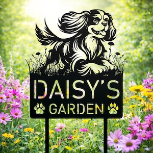 以下が含まれることがあります： 「DAISY'S GARDEN」の文字と足跡のシルエットが描かれた、キャバリア・キング・チャールズ・スパニエルのシルエットが特徴の黒い金属製のガーデンサイン。色とりどりの花が咲く庭に設置されています。