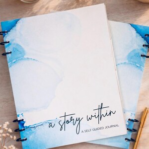 May include: Two spiral-bound journals with watercolor-style blue and white covers. The text "a story within" is written on the cover, along with "A SELF GUIDED JOURNAL". A gold pen rests beside the journals.