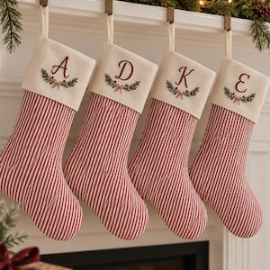 Peut inclure: Quatre chaussettes de Noël avec un motif à rayures verticales rouges et blanches sur la partie inférieure et un revers crème. Chaque revers est brodé d'une lettre, A, D, K ou E, au-dessus d'un motif de feuilles vertes et de baies rouges.