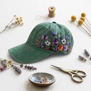 May include: A sage green baseball cap featuring detailed floral embroidery on the side. The design includes various small flowers in shades of purple, white, and orange, along with green stems and leaves, stitched directly onto the fabric surface.
