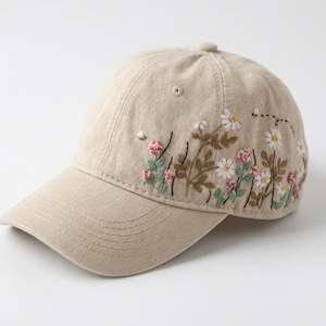 May include: A beige baseball cap featuring detailed embroidery of white daisies, pink clover flowers, and green foliage along the side. Small stitched bees are visible near the floral pattern, which is set against the light tan fabric of the cap.