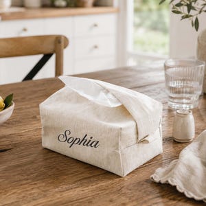 Peut inclure: Une housse de boîte à mouchoirs rectangulaire en tissu beige avec le prénom Sophia brodé en lettres cursives sombres sur le devant. La housse est fabriquée dans une matière texturée semblable à du lin et possède une ouverture sur le dessus.