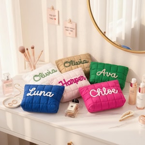 Op de afbeelding: Een verzameling gewatteerde make-up tasjes in verschillende kleuren, waaronder blauw, roze, groen en beige. Elke tas heeft een naam in een contrasterende kleur geborduurd. De tasjes liggen op een witte kaptafel met schoonheidsproducten.
