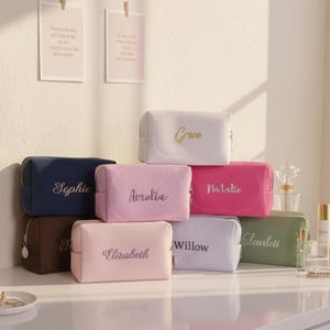 Könnte beinhalten: Eine Sammlung personalisierter Kosmetiktaschen in verschiedenen Farben, darunter Weiß, Rosa, Marineblau und Grün. Jede Tasche ist mit einem anderen Namen in einer passenden Farbe bestickt. Die Taschen sind auf einer weißen Oberfläche angeordnet, mit Schönheitsprodukten im Hintergrund.