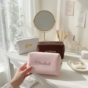 Può includere: Tre trousse per cosmetici personalizzate in bianco, marrone e rosa, ciascuna con un nome ricamato in un colore a contrasto. La borsa bianca dice "Grace" in oro, la borsa marrone dice "Willow" in bianco e la borsa rosa dice "Elizabeth" in rosa scuro.