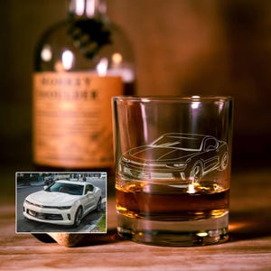 May include: A clear glass tumbler featuring a white etched line drawing of a Chevrolet Camaro sports car. The glass is filled with an amber-colored liquid. An inset image shows a white Chevrolet Camaro parked on a city street.