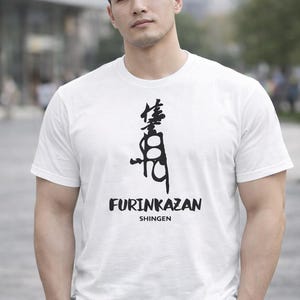 Puede incluir: Camiseta blanca con un diseño gráfico negro y el texto "FURINKAZAN SHINGEN". El diseño presenta caracteres japoneses encima del texto. La camiseta es de manga corta.