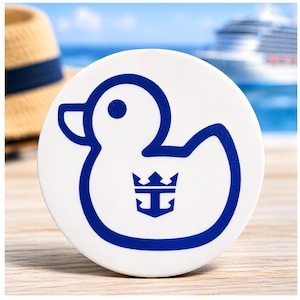 May include: A white, circular coaster featuring a blue outline of a duck with a crown and anchor emblem. A straw hat with a navy blue band is in the background, along with a cruise ship and ocean view.