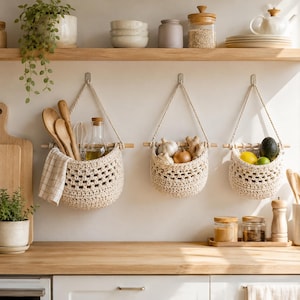 Puede incluir: Tres cestas colgantes de ganchillo en color blanco roto suspendidas de varillas de madera en una pared. Las cestas contienen cucharas de madera y un paño, ajo y jengibre, y fruta fresca que incluye un aguacate y un limón. Patrón texturizado.