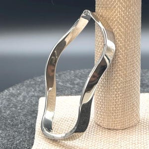 May include: A polished silver-tone bangle bracelet featuring a continuous, fluid wavy design. The metal surface is smooth and reflective, creating a series of gentle curves that form an irregular, organic shape throughout the entire band.