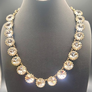 May include: A gold-toned necklace featuring a series of round, clear, faceted crystals set in metal prongs. The crystals are linked together in a continuous chain, creating a circular design that rests evenly around the neck area.