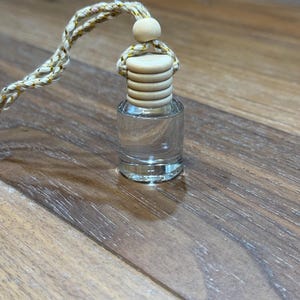 May include: A small, clear glass bottle with a wooden cap and a braided cord. The bottle is empty and appears to be for essential oils or fragrance. The cord is beige with gold accents.