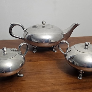 May include: A silver tea set comprising a teapot, a creamer, and a sugar bowl. The teapot features a curved spout and handle. The creamer and sugar bowl have handles and lids. All pieces are on small feet and rest on a wooden surface.