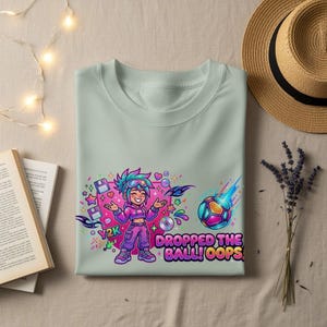 Puede incluir: Camiseta verde claro con un diseño gráfico colorido. El diseño presenta un personaje de dibujos animados, un balón de fútbol, corazones y estrellas, con el texto "Y2K DROPPED THE BALL! OOPS".