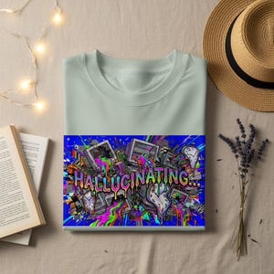 May include: A pale green crew neck t-shirt with a striking graphic. The design includes the word "HALLUCINATING" in a multi-coloured font, alongside melting clocks and computer monitors set against a blue backdrop.