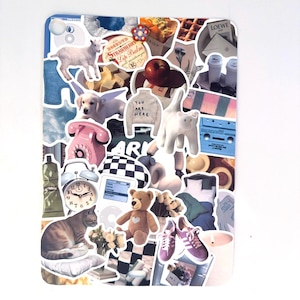 Puede incluir: Un collage decorativo de pegatinas con diversas imágenes. Incluye un teléfono rosa, un oso de peluche, un gato, una cabra, un cachorro alado, un suéter con el texto "YOU ARE HERE" y una cinta de cassette.