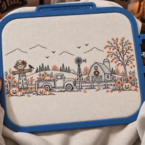 May include: A rectangular blue embroidery hoop holding a light-colored fabric panel with a black and orange line art design featuring a scarecrow, a pickup truck, a windmill, a barn, sunflowers, and pumpkins in a rural autumn landscape.