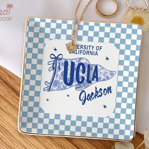 May include: Square ceramic trinket tray with a blue and white checkered pattern and gold trim. A white pennant with "UCLA Jackson" text is centered on the tray. A gold necklace and rings are on the tray.