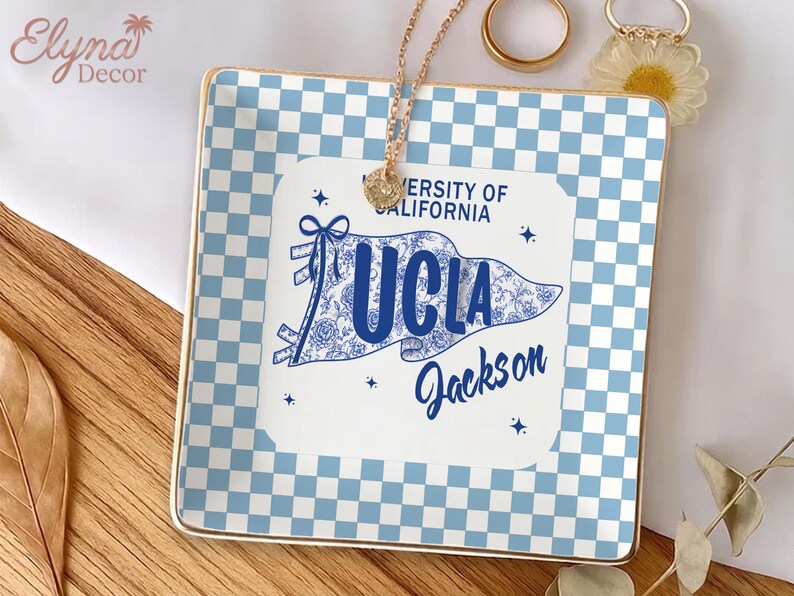May include: Square ceramic trinket tray with a blue and white checkered pattern and gold trim. A white pennant with "UCLA Jackson" text is centered on the tray. A gold necklace and rings are on the tray.