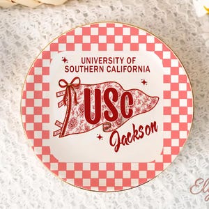 May include: A decorative plate with a red and white checkered border and a gold rim. The centre features the text "UNIVERSITY OF SOUTHERN CALIFORNIA" and a banner with "USC" and "Jackson" in red script. The banner has a floral design.