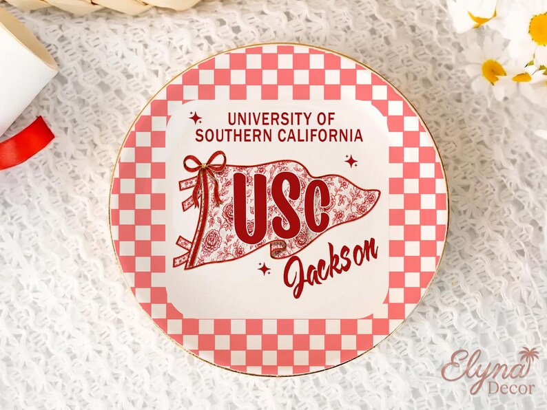 May include: A decorative plate with a red and white checkered border and a gold rim. The centre features the text "UNIVERSITY OF SOUTHERN CALIFORNIA" and a banner with "USC" and "Jackson" in red script. The banner has a floral design.