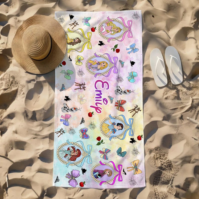 May include: A rectangular beach towel featuring a pastel watercolor background with various cartoon princess illustrations in oval frames, bows, apples, and the name Emily written in purple script, accompanied by a straw hat and white flip-flops.
