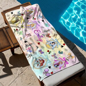 May include: A rectangular beach towel featuring a pastel watercolor background with the name Emily printed in purple script. The towel displays various Disney princess illustrations in ornate frames, colorful bows, red roses, and small castle icons.