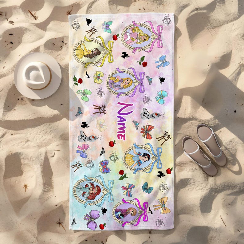 May include: A rectangular beach towel with a watercolor pastel background featuring various cartoon princess characters in ornate oval frames, small bows, red apples, and the word Name printed in purple. The towel is displayed on a sandy surface.