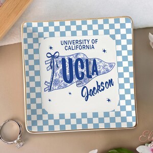May include: A square ceramic trinket tray with a blue and white chequered pattern and gold trim. The centre features a UCLA pennant design with the text "University of California UCLA Jackson". A silver ring is in the foreground.