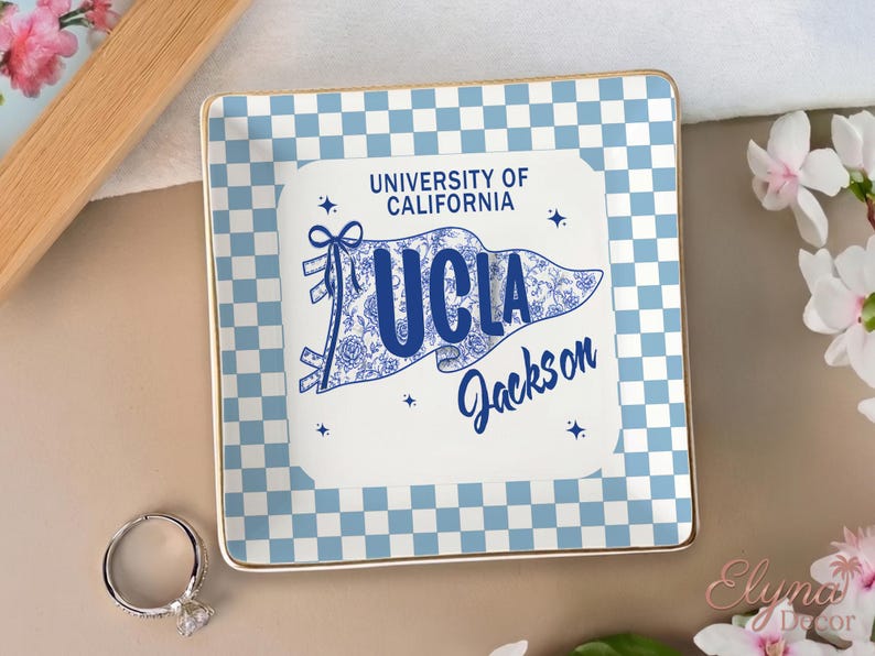 May include: A square ceramic trinket tray with a blue and white chequered pattern and gold trim. The centre features a UCLA pennant design with the text "University of California UCLA Jackson". A silver ring is in the foreground.
