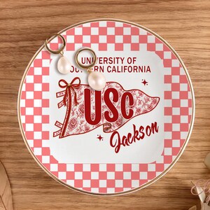 May include: A decorative ceramic plate with a gold rim, featuring a red and white checkered pattern and a central design with the text "UNIVERSITY OF SOUTHERN CALIFORNIA" and "USC Jackson". Two pearl earrings rest on the plate.