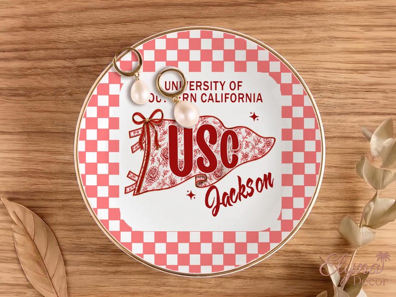 May include: A decorative ceramic plate with a gold rim, featuring a red and white checkered pattern and a central design with the text "UNIVERSITY OF SOUTHERN CALIFORNIA" and "USC Jackson". Two pearl earrings rest on the plate.