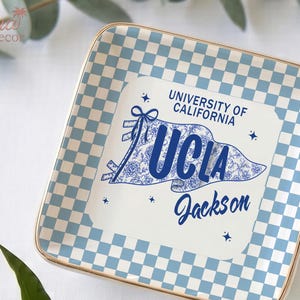 May include: Square ceramic tray with a light blue and white chequered pattern border and a gold rim. The centre features a UCLA banner design with the text "UNIVERSITY OF CALIFORNIA UCLA Jackson" in blue.