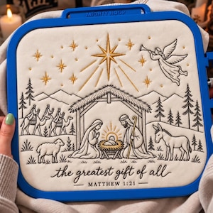 Könnte beinhalten: Ein quadratischer blauer Mighty Hoop Stickrahmen mit cremefarbenem Stoff, der eine Krippenszene in Schwarz und Gold zeigt. Das Design umfasst einen Stern, einen Engel, die heilige Familie, Tiere sowie den Text "the greatest gift of all" und "MATTHEW 1:21".