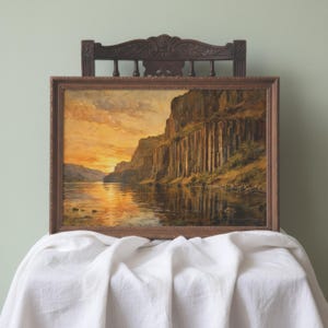 May include: A framed landscape painting depicting a river flowing past tall, vertical basalt cliffs at sunset. The sky is filled with warm orange and yellow hues, which are reflected in the calm water below. The frame is a dark, textured wood.