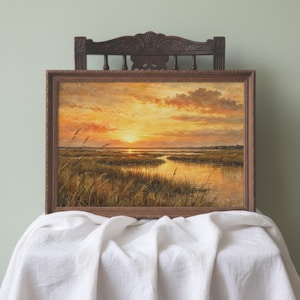 May include: A landscape painting in a dark brown wooden frame depicting a sunset over a marshland. The scene features golden and orange clouds above a reflective waterway surrounded by tall grasses, captured in a horizontal rectangular orientation.