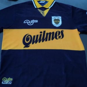 May include: A navy blue and yellow soccer jersey with the word "Quilmes" across the chest. The jersey has a yellow horizontal stripe across the chest and a yellow collar. The team logo is on the chest.