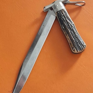 May include: A stainless steel folding knife with a long blade, a textured brown handle, and a silver metal bolster. The knife is partially open, displaying the blade against an orange background.