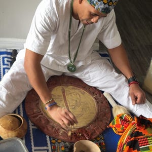 Puede incluir: Una persona con camisa y pantalón blancos y un pañuelo estampado en la cabeza, sentada sobre una esterilla azul. Dibuja con el dedo sobre una tabla de madera circular con centro dorado, rodeada por un cuenco con piedras oscuras y un recipiente de calabaza.