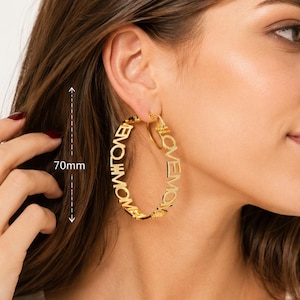 May include: Gold hoop earrings with the words "LOVE MOM" in a script font. The earrings are approximately 7 cm in diameter. The earrings are worn on a person.