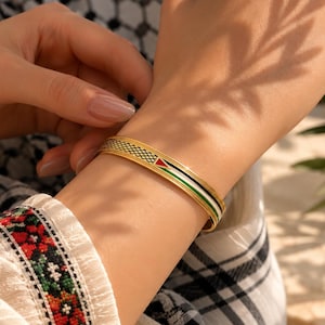 Pode incluir: Uma pulseira de metal dourado com um padrão de grade preto texturizado de um lado e um design de listras horizontais nas cores esmaltadas vermelho, preto, branco e verde. A pulseira tem um acabamento polido e um design fino e aberto.