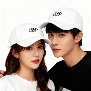 May include: Two white baseball caps with the word "GRAP" embroidered in blue. The caps are worn by a person. The caps have a curved brim and an adjustable strap at the back. The background is white.