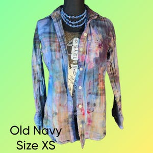 May include: A button-down, long-sleeve shirt with a colorful tie-dye pattern in shades of blue, purple, and pink. The shirt is layered over a graphic tee and accessorized with a blue beaded necklace. The text "Old Navy Size XS" is visible.