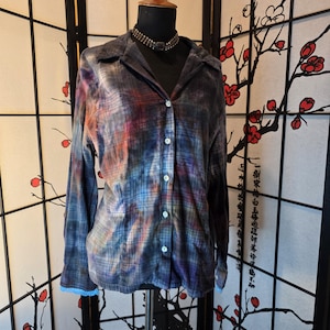 May include: A button-down shirt with a plaid pattern in shades of blue, purple, red, and orange. The long-sleeved shirt has a collar and is displayed on a mannequin. The cuffs have a light blue lace trim.