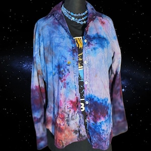 Pode incluir: Uma camisa abotoada listrada em azul e branco com detalhes tie-dye em tons de azul, roxo e vermelho. A camisa é sobreposta a uma camiseta preta com uma estampa gráfica. Um colar de contas azuis também é visível.
