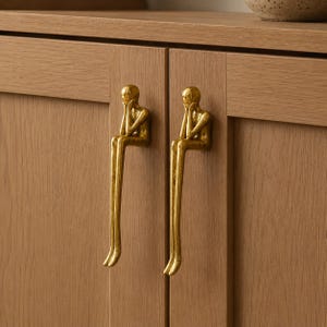 Peut inclure: Deux poignées de placard dorées, chacune en forme de personne assise, sont montées sur une armoire en bois brun clair. Les poignées ont un design artistique unique, ajoutant un élément décoratif aux portes de l'armoire.