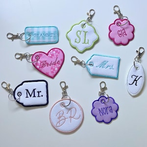Peut inclure: Une collection de neuf porte-clés en tissu brodé de formes variées, notamment des rectangles, des cœurs, des cercles et des fleurs festonnées. Chaque porte-clés comporte un mousqueton en métal et un texte ou des initiales personnalisés en bleu, rose, blanc et violet.