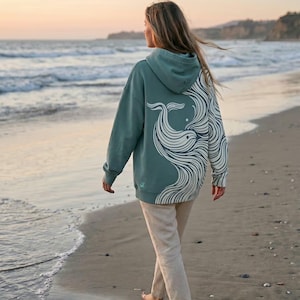 May include: A teal hoodie with a white whale design on the back. The hoodie is worn by a person walking on a sandy beach near the ocean. The design features a whale with flowing lines.