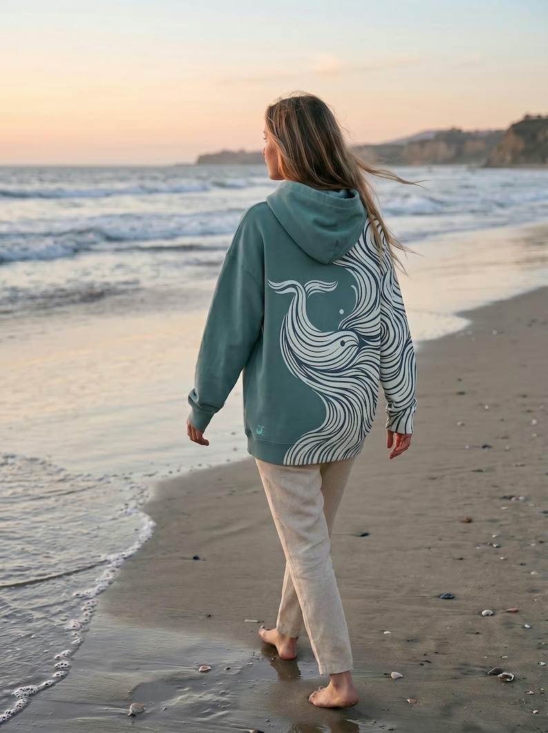 May include: A teal hoodie with a white whale design on the back. The hoodie is worn by a person walking on a sandy beach near the ocean. The design features a whale with flowing lines.