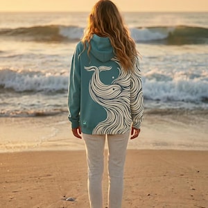 May include: A teal hoodie with a whale design on the back. The whale is depicted in white, flowing lines. The person is standing on a sandy beach, facing the ocean at sunset. The hoodie has a hood and long sleeves.